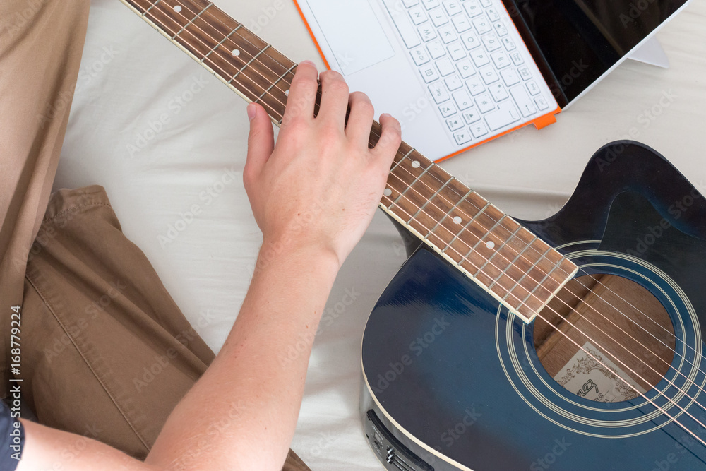 Fototapeta premium Junger Mann sitzend im Bett, Gitarre und Laptop