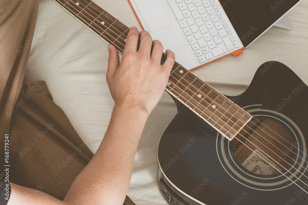 Fototapeta premium Junger Mann sitzend im Bett, Gitarre und Laptop