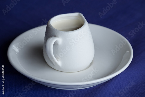 Fototapeta Naklejka Na Ścianę i Meble -  White ceramic creamer on a saucer with cold cream on a blue background.