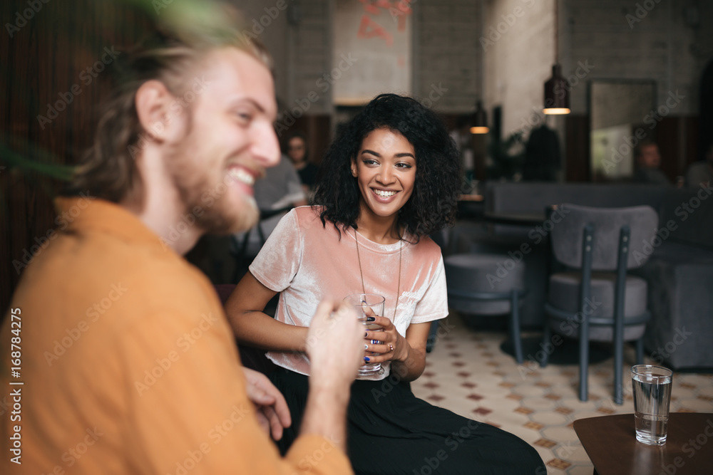 Beautiful smiling girl sitting in restaurant with friend. Joyful African American lady sitting at cafe with glass of water in hand. Young girl with dark curly hair talking with friend at cafe