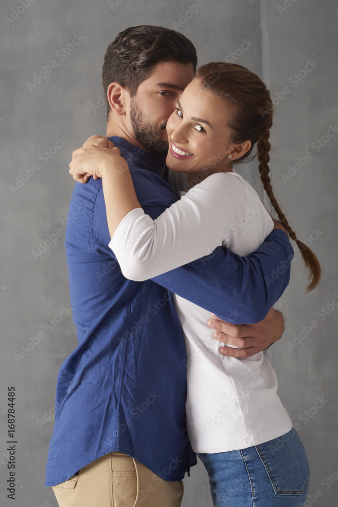 Enjoy every moment together. Studio portrait of a loving young couple embracing each other while standing at grey wall and looking at camera. Enjoy every moment together. Studio portrait of a loving young couple embracing each other while standing at grey wall and looking at camera.