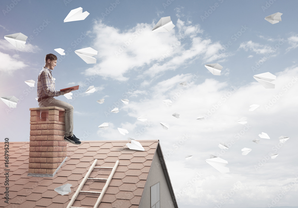 Man on brick roof reading book and paper planes flying in air Man on brick roof reading book and paper planes flying in air