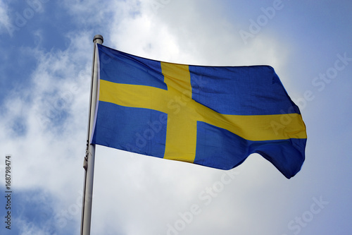 Flag of Sweden with a yellow cross on a blue background, national symbol or sign of the european country, fluttering in the wind against the blue sky with clouds on a sunny day
