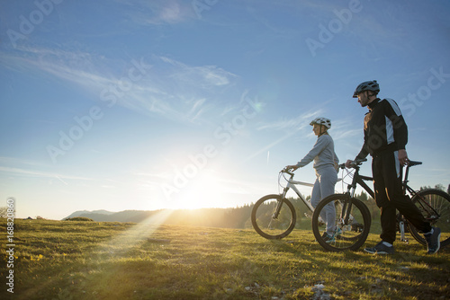 Cyclist couple with mountain bikes standing on the hill under the evening sky and enjoying bright sun at the sunset. © FS-Stock