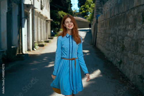 Beautiful young woman in blue dress with red hair walking along the boulevard in the city