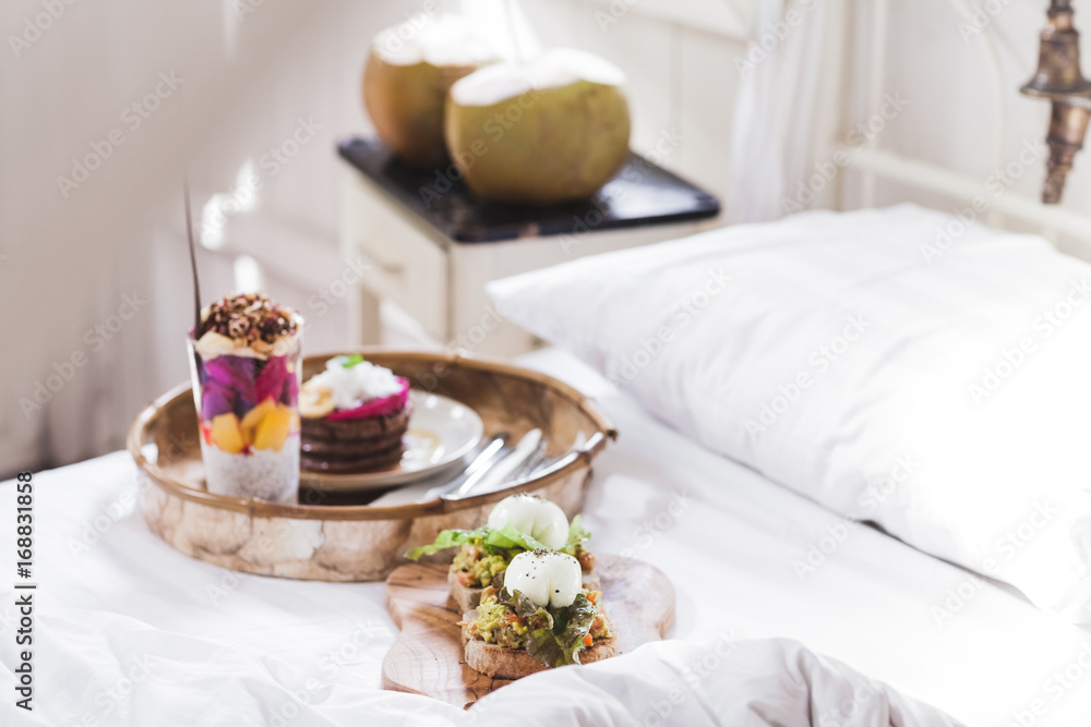 Fresh tasty breakfast in bed on wooden tray. Yogurt with fruits and chia, honey pancakes, two toasts with avocado and poached eggs and two young coconut. White vintage interior Fresh tasty breakfast in bed on wooden tray. Yogurt with fruits and chia, honey pancakes, two toasts with avocado and poached eggs and two young coconut. White vintage interior
