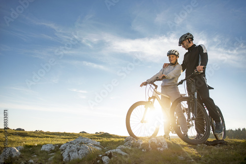 Fototapeta Naklejka Na Ścianę i Meble -  Cyclist couple with mountain bikes standing on the hill under the evening sky and enjoying bright sun at the sunset.