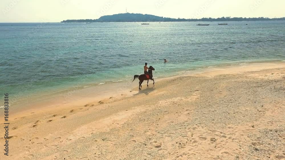 Aerial view of indonesian man riding black horse on beach. Shot with drone on sunny day with blue sky in Gili Meno, Indonesia. Aerial view of indonesian man riding black horse on beach. Shot with drone on sunny day with blue sky in Gili Meno, Indonesia.