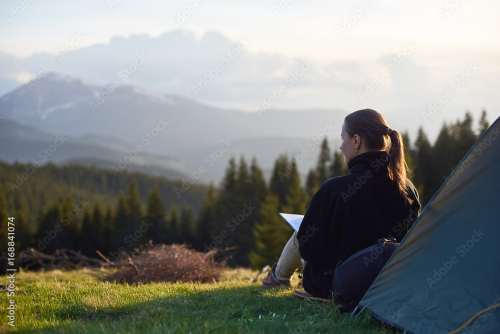 Fototapeta premium Rear view of young woman enjoying sunset in the mountains with a book near the tent