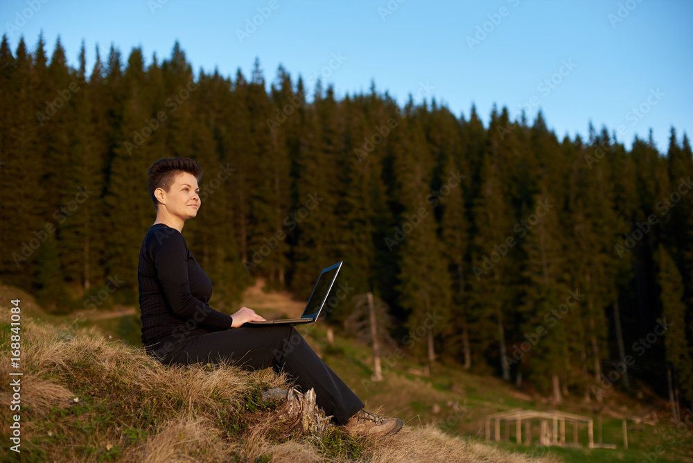 Joyful young attractive female working on her laptop outdoors enjoying view of the mountains copyspace Joyful young attractive female working on her laptop outdoors enjoying view of the mountains copyspace