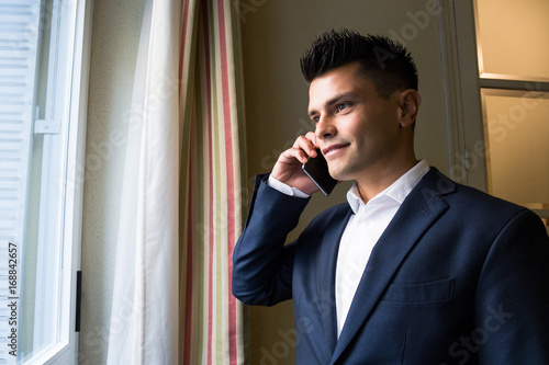 Young confident businessman wearing suit and talking smartphone looking smilingly away near window photo