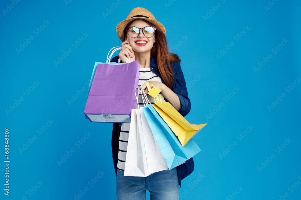 Beautiful woman holds many packages on a blue background in a hat and glasses, smile, shopping, shopper, shop, shopaholic Beautiful woman holds many packages on a blue background in a hat and glasses, smile, shopping, shopper, shop, shopaholic
