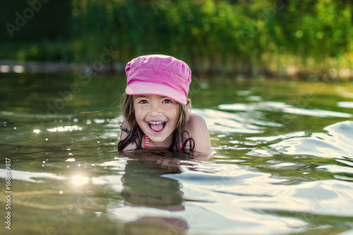 Fototapeta Naklejka Na Ścianę i Meble -  Happy kid girl in pink cap having fun time and swimming in lake water. Concept of summer vacation