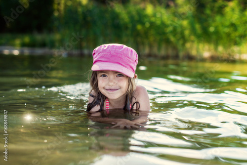 Fototapeta Naklejka Na Ścianę i Meble -  Smiling kid girl in pink cap having fun time and swimming in lake water. Concept of summer vacation