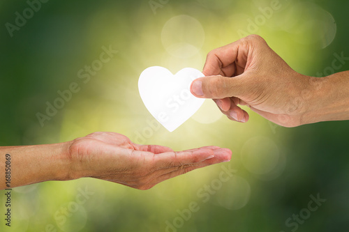 Hand holding and giving white heart to receiving hand on blurred green bokeh background, helping hand concept
