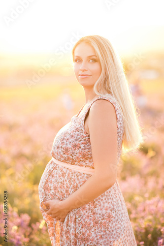 Beautiful blonde pregnant woman holding her tummy in meadow. Wearing stylish dress outdoors. Looking at camera. Maternity. Motherhood.