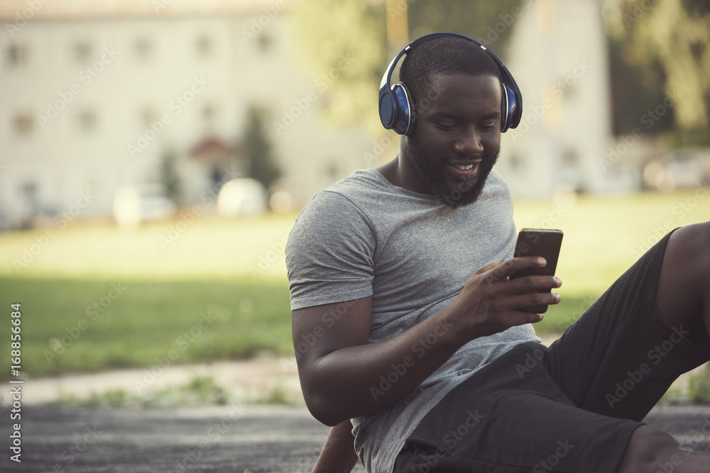 A handsome black man runner is resting on the track while using an app in his mobile smart phone to listen to music, wearing headphones A handsome black man runner is resting on the track while using an app in his mobile smart phone to listen to music, wearing headphones