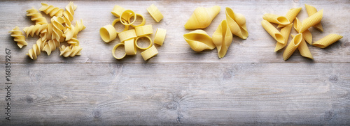 Raw pasta: fusilli, paccheri, conchiglie, and penne on wooden background. Top view, space for text.