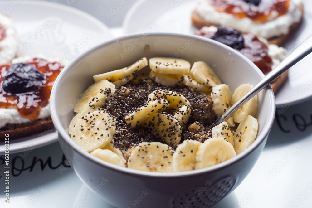 White bowl of cornflakes slice of banana and chia seeds, dark bread with white cheese and jam in background
