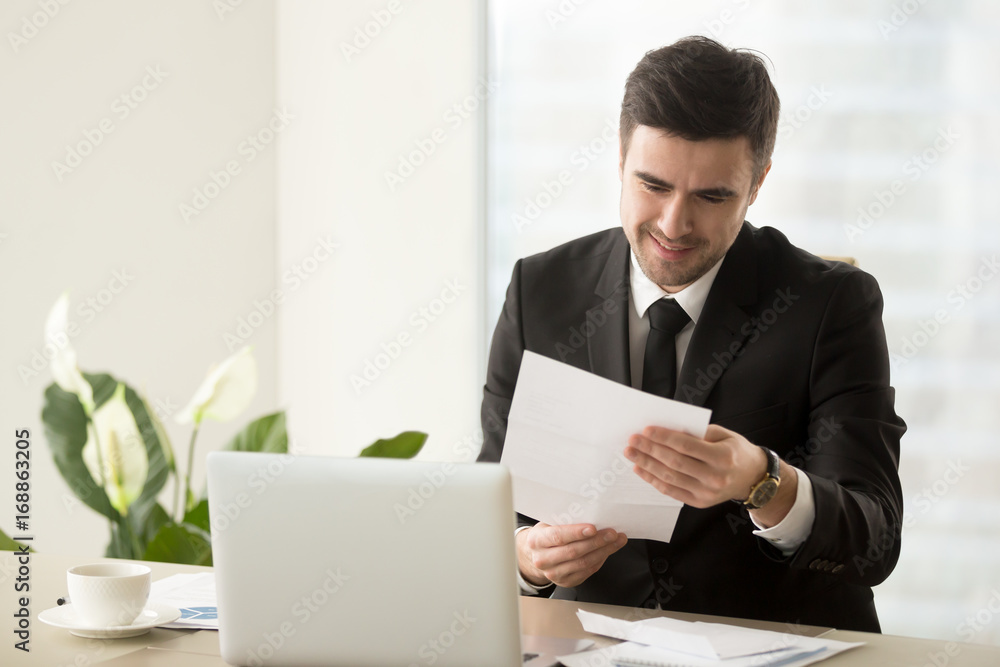Smiling successful businessman holding document sitting at desk, reading good news in letter, reviewing beneficial lucrative contract, getting notification about bank loan or business credit approval Smiling successful businessman holding document sitting at desk, reading good news in letter, reviewing beneficial lucrative contract, getting notification about bank loan or business credit approval
