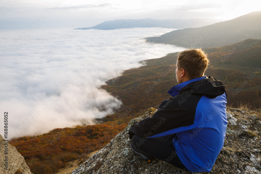 Young tourist hiker with backpack siting and relaxing on the top of the mountain and looking at beautiful yellow autumn landscape sunset over clouds