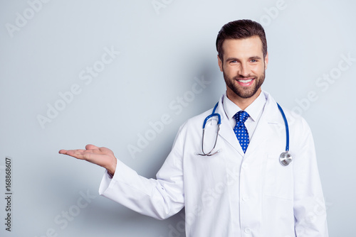 Cheerful handsome bearded smiling guy is holding the copy space. Doc is wearing white uniform and a tie, stands on a light white background