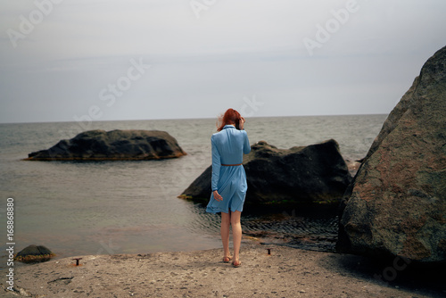 Young woman in a blue dress is standing on the beach near the rocks