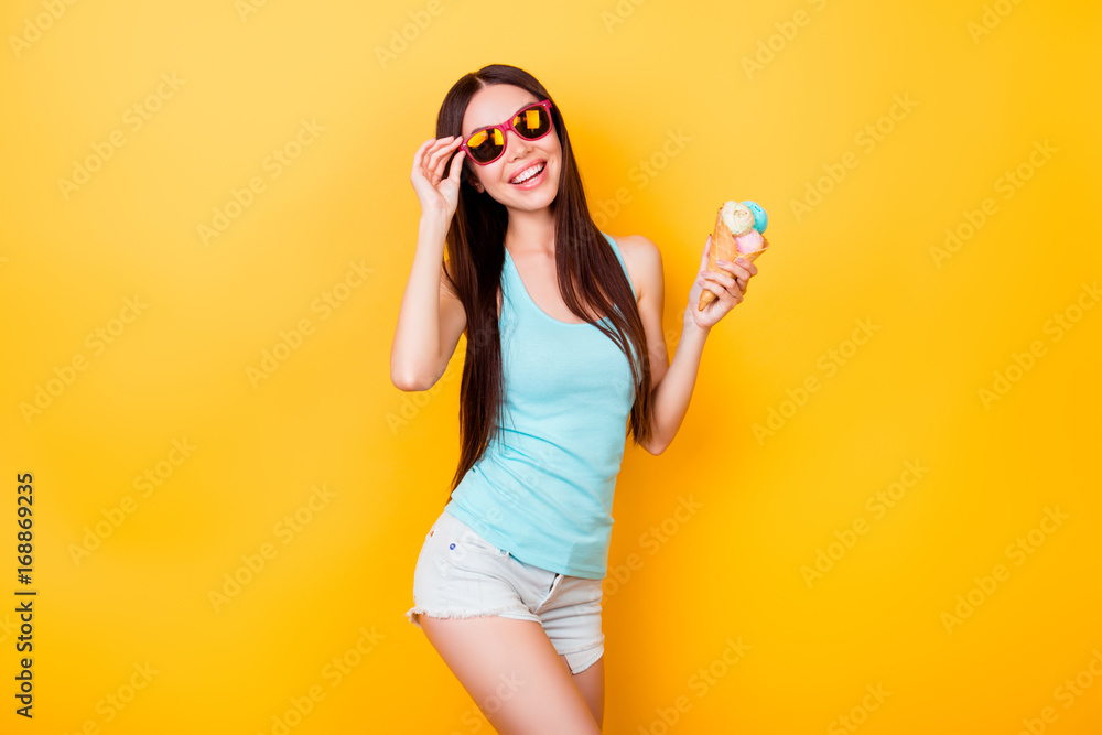 Glad delightful young asian woman with toothy smile looks at camera, holds tasty ice cream of three scoops, stands on yellow background in tourist outfit, glasses Glad delightful young asian woman with toothy smile looks at camera, holds tasty ice cream of three scoops, stands on yellow background in tourist outfit, glasses