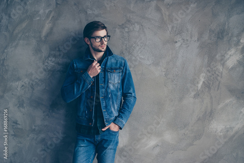 Stylish young handsome guy in a denim suit stands on a gray background and looks to the side