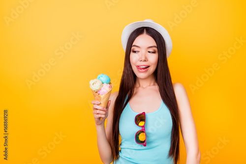 Glad delightful young asian woman with tongue out looks at tasty ice cream of three scoops of different flavors, stands on yellow background in tourist outfit © deagreez