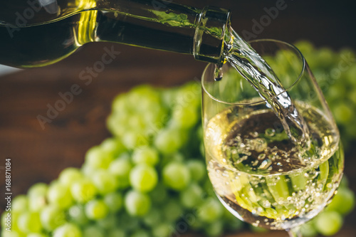 Pouring white wine into a glass with a bunch of green grapes against wooden background