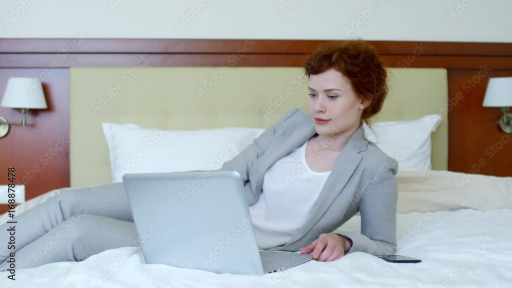 Elegant young business lady in formal suit lying on bed in hotel room and typing on laptop Elegant young business lady in formal suit lying on bed in hotel room and typing on laptop