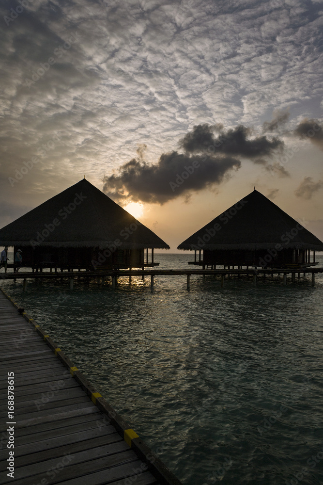 Dramatic sky at sunset and silhouettes of two bungalows overlooking the island of Maledives
