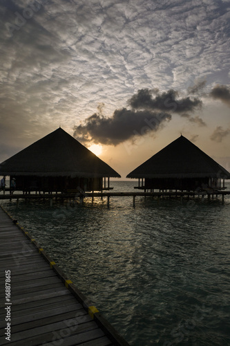 Dramatic sky at sunset and silhouettes of two bungalows overlooking the island of Maledives