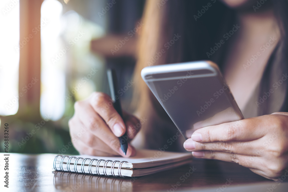 Closeup image of a business woman writing on notebook while using and holding mobile phone on wooden table background in office Closeup image of a business woman writing on notebook while using and holding mobile phone on wooden table background in office