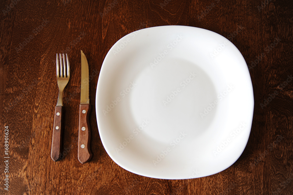 Empty white plate with a fork and knife on a dark wooden brown table Empty white plate with a fork and knife on a dark wooden brown table