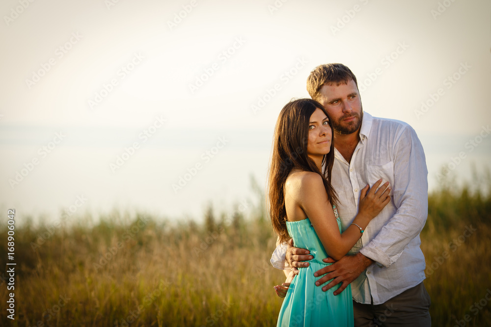 A young and beautiful woman put her hand on her husband's torso, a loving couple stands in the grass near the sea, on the street a warm and summer day