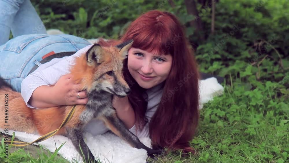 Young woman lies on grass in park with red fox outdoors. She gently caresses fluffy wool of cute pet with leash around her neck, pleasantly spending time in verdant parkland on summer day. Young woman lies on grass in park with red fox outdoors. She gently caresses fluffy wool of cute pet with leash around her neck, pleasantly spending time in verdant parkland on summer day.