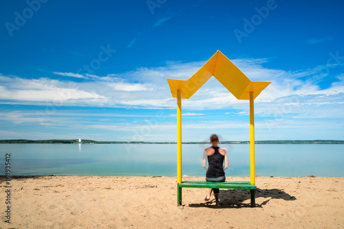 Silhouette of a girl sitting on a yellow metal bench with a canopy from the sun on the beach. Relaxation and recreation concept. Long exposure © v_sot