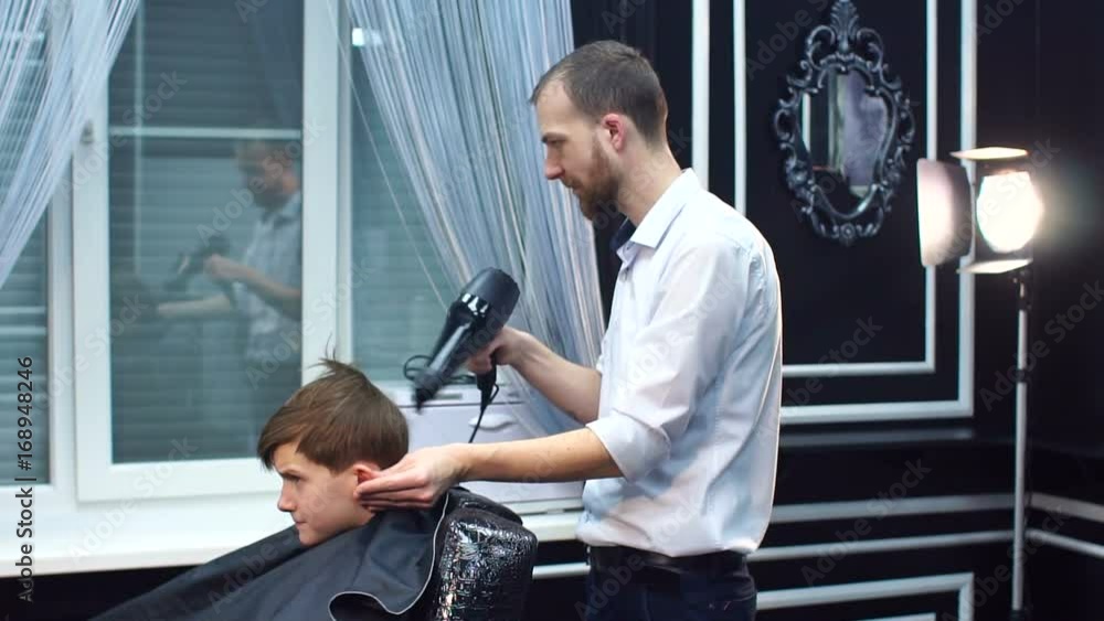 Modern bearded hairstylist drying hair with Hairdryer to teenager in a trendy barbershop. Modern bearded hairstylist drying hair with Hairdryer to teenager in a trendy barbershop.