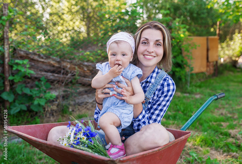Mother and her little daughter sit in the wheelbarrow outdoors