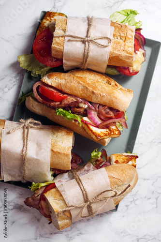 Fresh baguette sandwich bahn-mi styled. Bacon, roasted cheese, tomatoes and lettuce on metallic tray on white marble background.