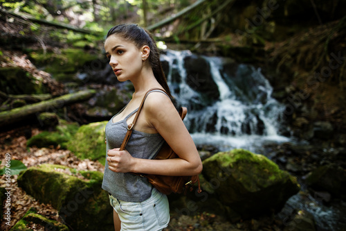 Hiking in forest with waterfall in background. Woman trekking in mountains against waterfall. Female with backpack on hike in nature. Cascading waterfall