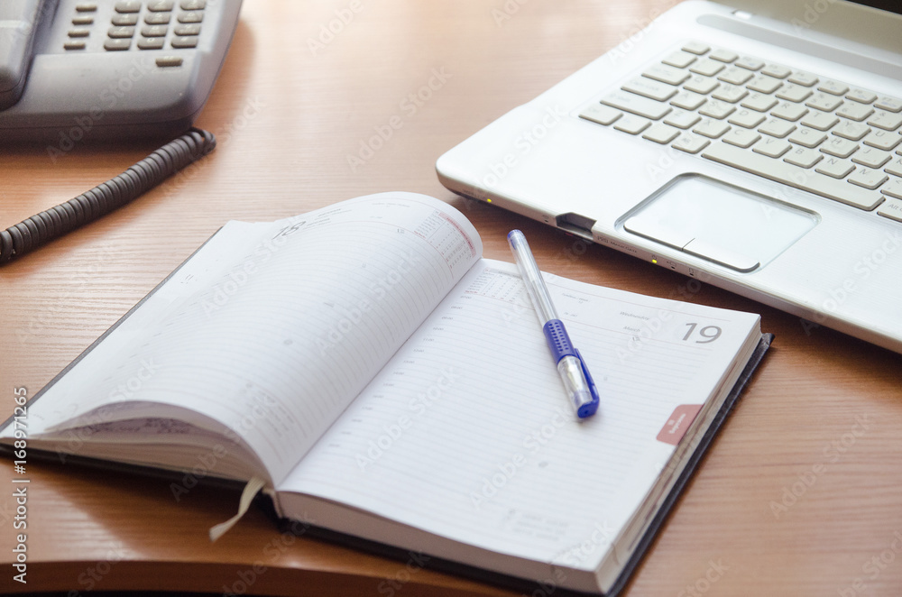 On the table is a laptop, a detailed notebook with pen and landline phone On the table is a laptop, a detailed notebook with pen and landline phone