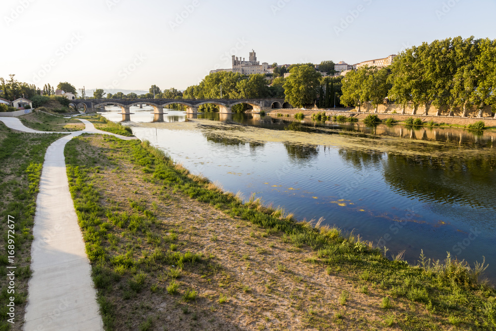 Views at sunset of the French city of Beziers, with trees and one of its bridges reflected over the river Orb, and the 13th-century Cathedral of Saint Nazaire in the background