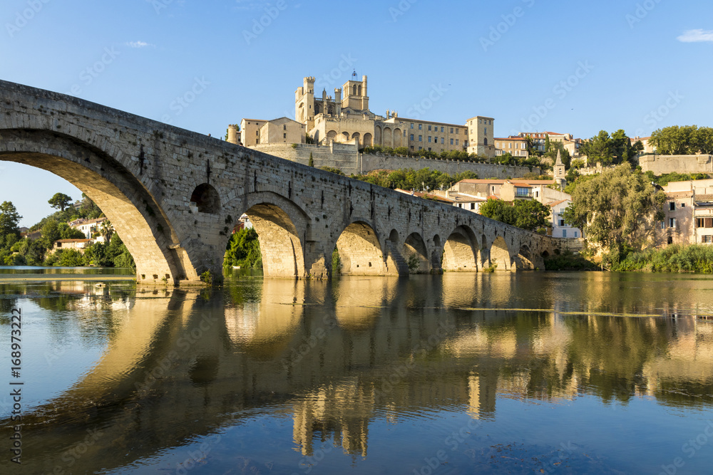 Views at sunset of the French city of Beziers, with trees and the old bridge reflected over the river Orb, and the 13th-century Cathedral of Saint Nazaire in the background