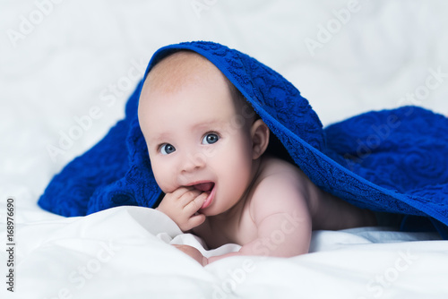 Cute smiling baby girl or boy after shower with towel on head in white sunny bedroom. Child with big blue eyes chews his hand and looks at the camera