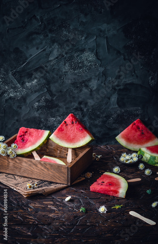 watermelon slices on wooden sticks in wooden box with white flowers on brown wooden table on dark blue background
