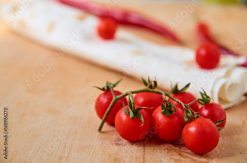 Branch of red cherry tomatoes isolated on light brown wooden background close up. On blurred background red chili pepper, tomatoes and pita. Side view