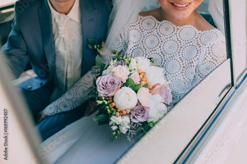 Beautiful bride in a white dress and groom in tuxedo are sitting in a car and holding a wedding bouquet. Groom embrace woman by the waist. Artwork